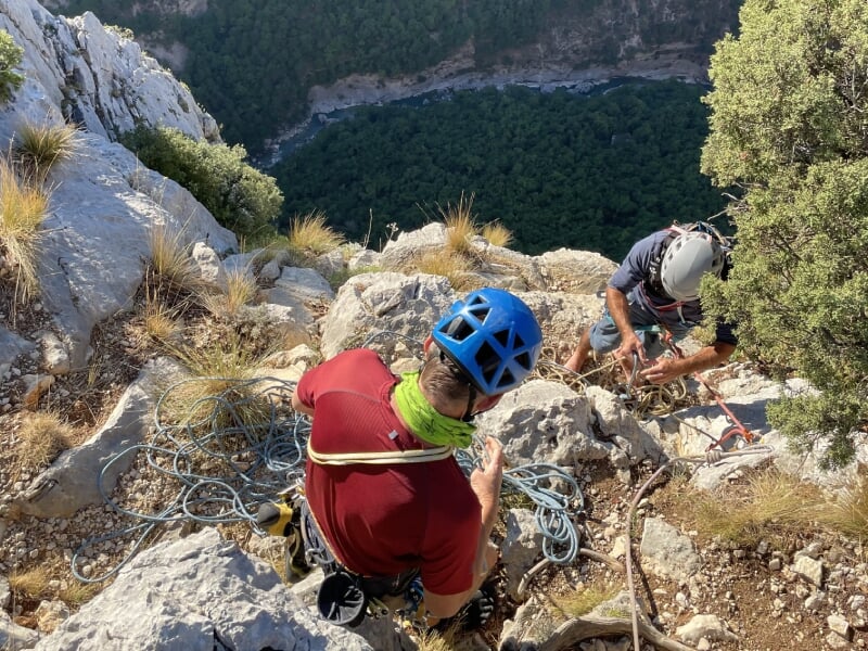 Deux alpinistes, l'un en casque bleu et l'autre en casque blanc, organisant des cordes sur un sentier rocheux, entourés de végétation.