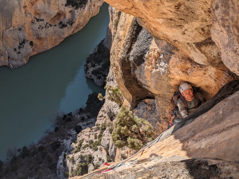 Escalade difficile dans une paroi des Gorges du Verdon, grimpeur souriant équipé d'un casque et de cordes, vue plongeante sur une rivière.