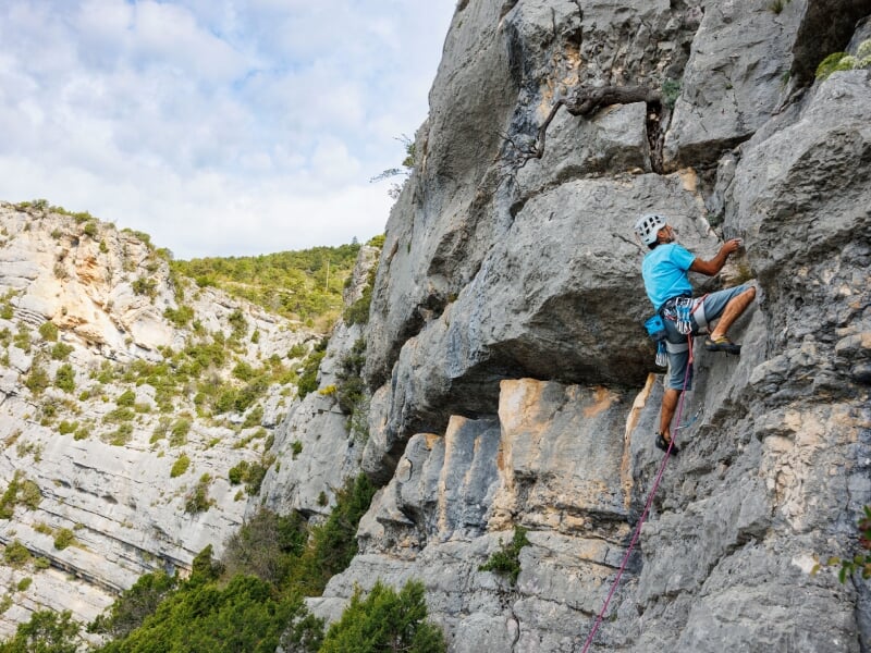 Grimpeur en t-shirt bleu et casque d'escalade, progressant sur une falaise rocheuse, sécurisé par une corde, entouré d'un paysage de montagnes et de végétation sous un ciel clair.