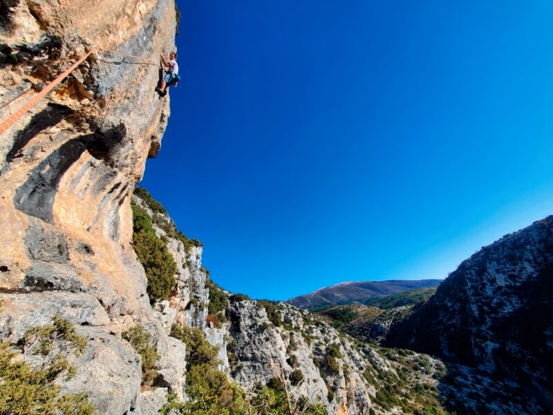 Grimpeur sur une haute paroi rocheuse, vêtu d'un t-shirt blanc et équipé d'un harnais, escaladant dans un cadre de montagnes escarpées et ciel bleu éclatant.