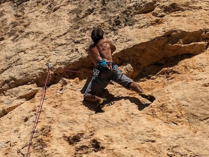 Grimpeur torse nu s'accrochant à une paroi rocheuse, vêtu d'un short et équipé d'un harnais et d'une corde de sécurité, démontrant force et endurance en escalade.