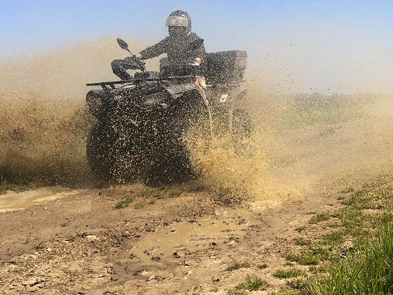 Un homme portant un casque et une combinaison noirs passe en quad dans une flaque en soulevant une gerbe d'eau