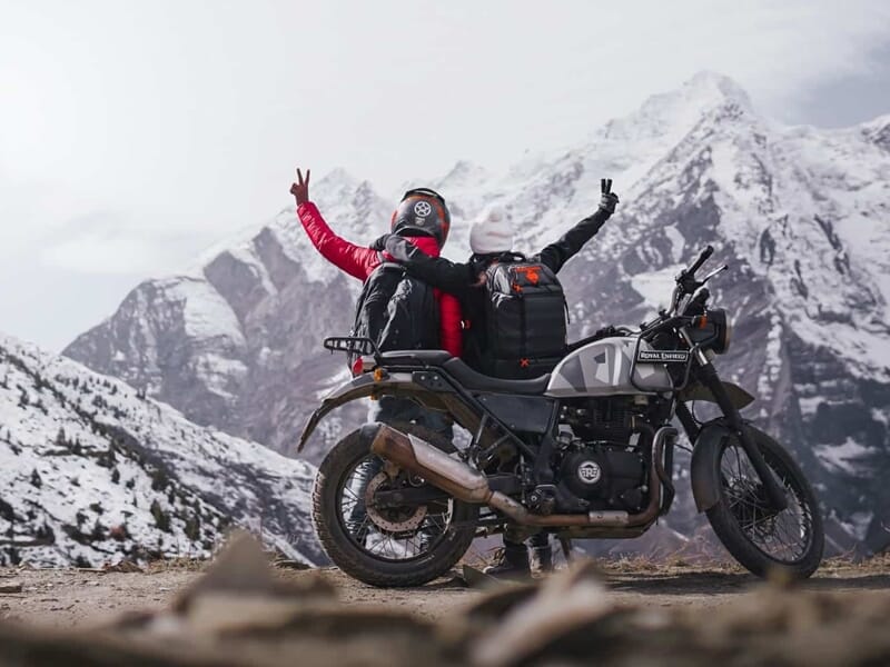 Couple de motards levant les bras en signe de victoire sur un parking devant un magnifique paysage de montagnes couvertes de neige