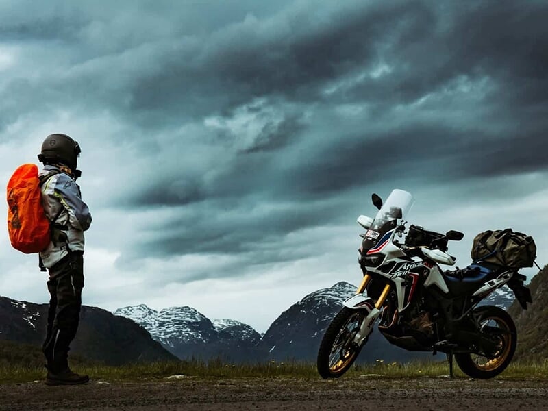 Motard à l'arrêt devant sa moto avec un paysage de montagnes enneigées en toile de fond