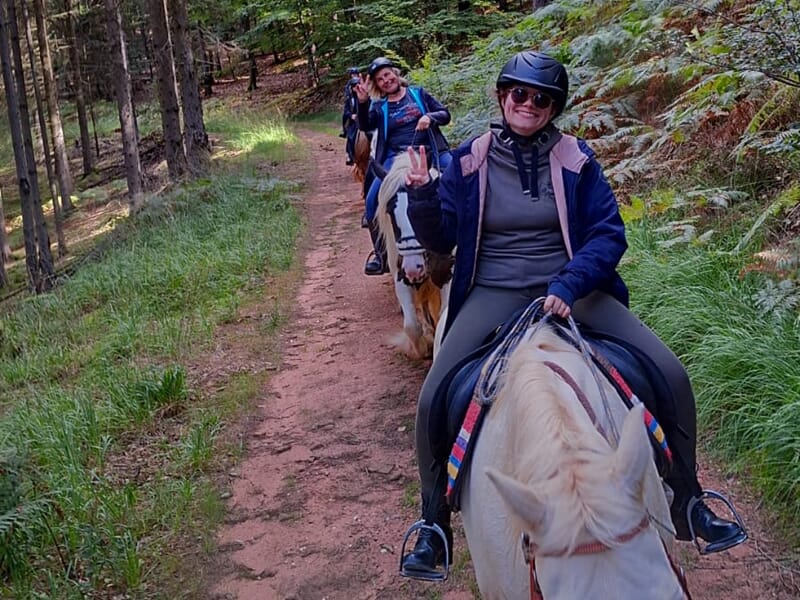 Cavalière souriante en tête d’un groupe sur un sentier forestier, faisant le signe de la paix en regardant l’objectif.