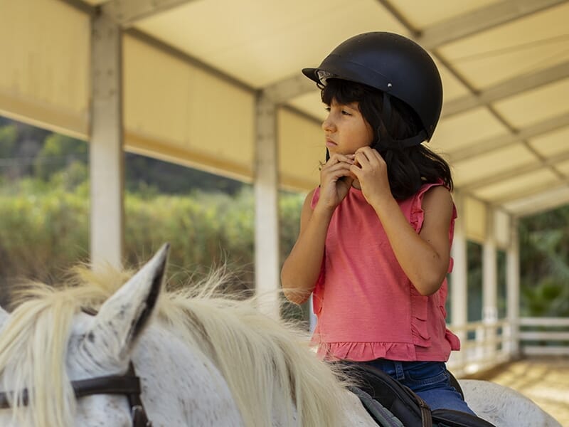 Jeune fille en chemise rose ajustant son casque d'équitation, montée sur un cheval blanc dans une zone couverte.