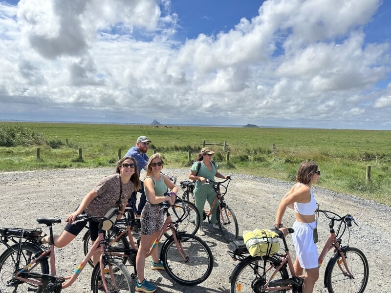 Cinq cyclistes posent souriants devant le Mont Saint-Michel sous un ciel partiellement nuageux, profitant d'une sortie en plein air.