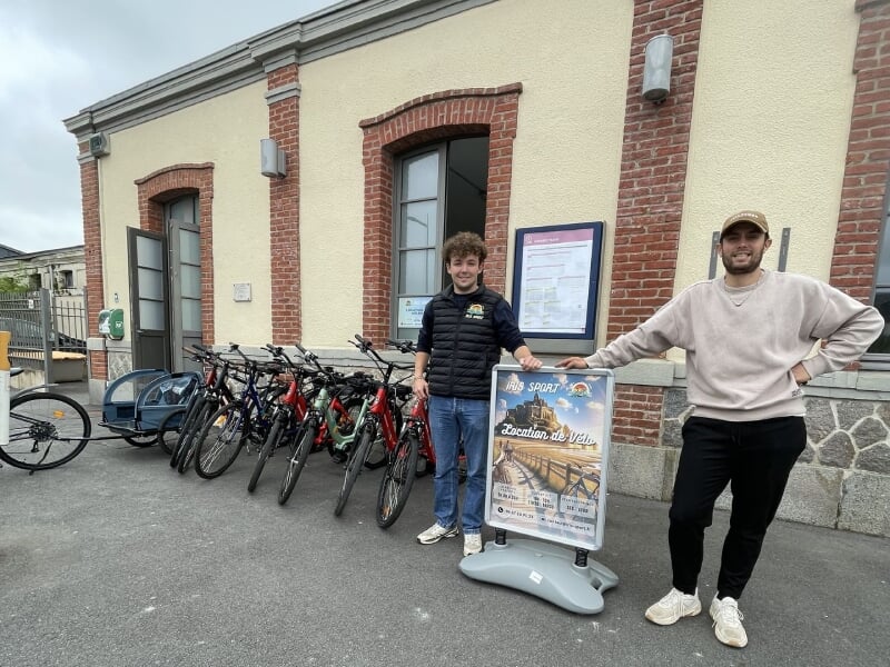 Deux hommes souriants devant un magasin de location de vélos avec plusieurs bicyclettes alignées et un panneau publicitaire.
