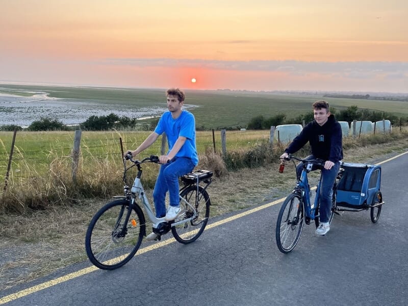 Deux hommes à vélo sur une route de campagne, avec un magnifique coucher de soleil en arrière-plan. L'un porte un t-shirt bleu, l'autre un sweat noir avec une remorque bleue.