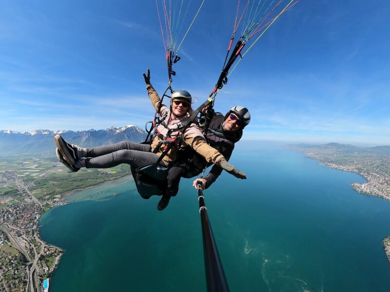 Parapentistes en tandem survolant un lac, vue plongeante sur une ville et des montagnes enneigées à l'arrière-plan, ciel bleu dégagé.