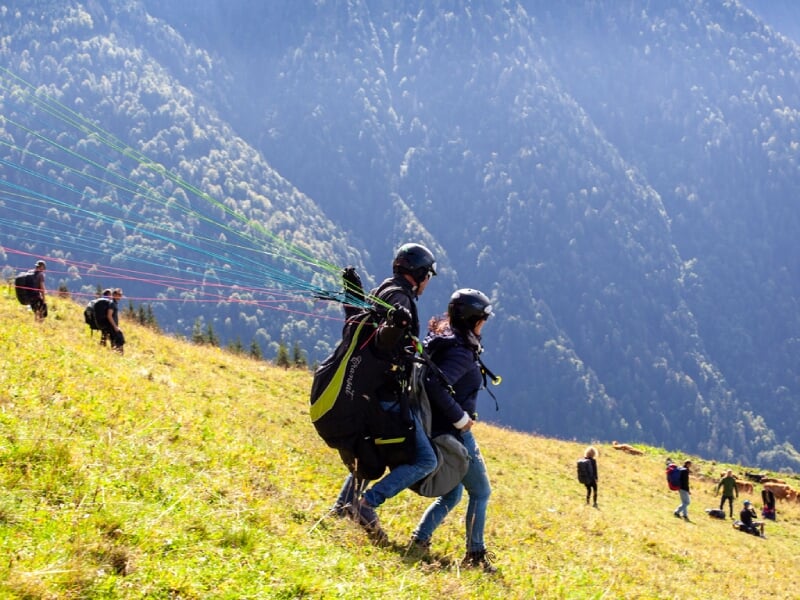 Décollage de parapente en tandem sur une pente herbeuse, forêt dense et autres parapentistes en arrière-plan.