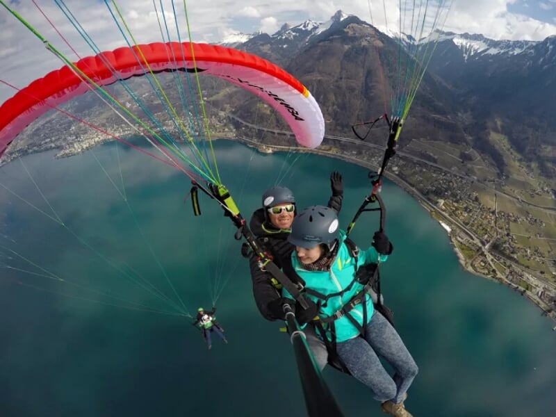 Parapentistes en tandem avec voile rouge survolant un lac, montagnes enneigées et zones résidentielles visibles en contrebas.