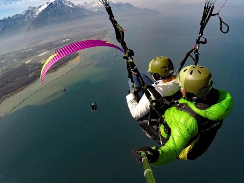Parapentistes en tandem avec voile rose survolant un lac, montagnes et littoral en arrière-plan.