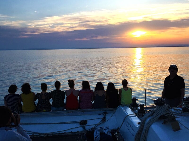 Groupe de personnes assises à l'arrière d'un voilier, contemplant un coucher de soleil sur une mer calme. Les reflets du soleil se mêlent aux teintes orangées du ciel