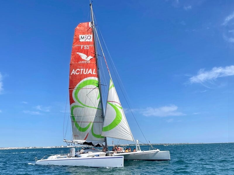 Trimaran avec une grande voile rouge et blanche naviguant sur une mer calme, sous un ciel bleu. Le bateau porte le nom 