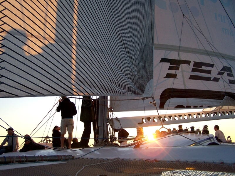 L'équipage d'un voilier manœuvrant les voiles au coucher du soleil, avec des ombres projetées sur la toile et une lumière dorée illuminant le pont