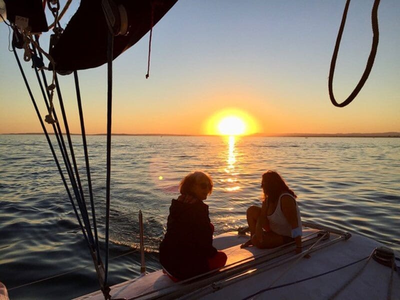 Deux personnes assises à l'avant d'un voilier au coucher du soleil, discutant tranquillement alors que les rayons du soleil disparaissent à l'horizon