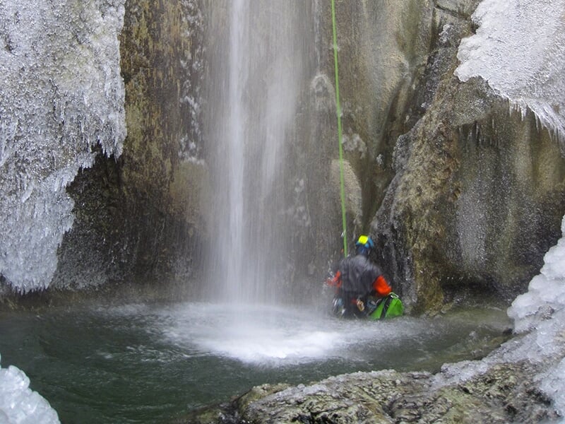 Un canyoneur en combinaison néoprène se tient debout dans la vasque d'une cascade avec de l'eau jusqu'à la taille