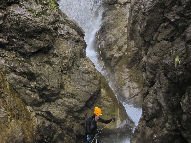Un canyoneur portant un casque orange termine une descente en rappel au pied d'une cascade tumultueuse