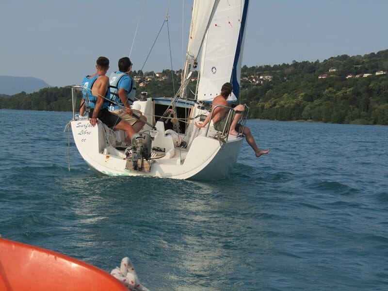 Trois plaisanciers de dos à bord d'un voilier sur un lac avec des collines en arrière-plan