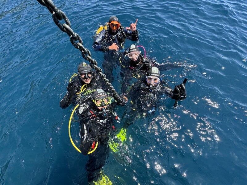 Groupe de plongeurs en combinaison noire à la surface de l'eau, souriant et faisant des signes de la main, à côté d'une chaîne de bateau