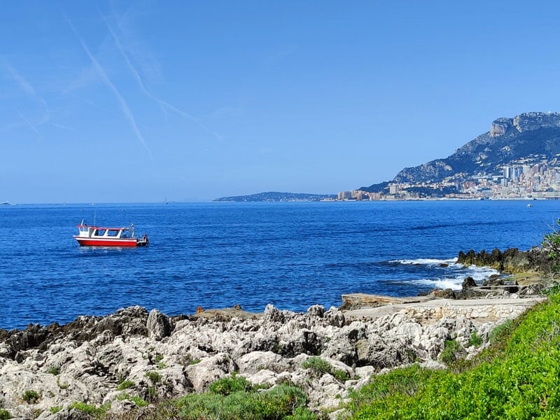 Bateau de pêche rouge et blanc flottant sur une mer calme, avec une ville et des montagnes en arrière-plan sous un ciel bleu clair