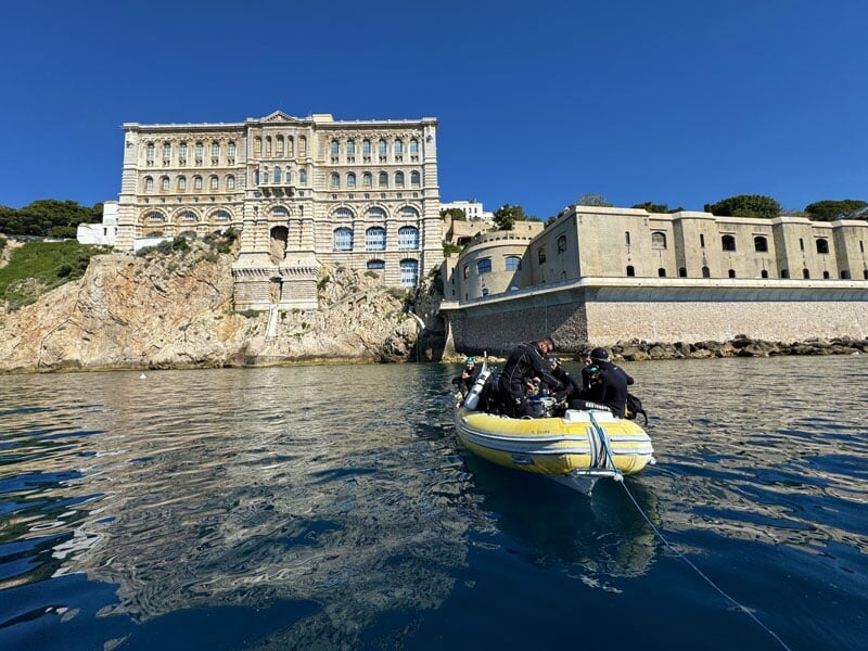 Petit bateau gonflable jaune amarré devant un grand bâtiment en pierre, avec plusieurs plongeurs à bord, prêt à plonger dans une eau calme et bleue