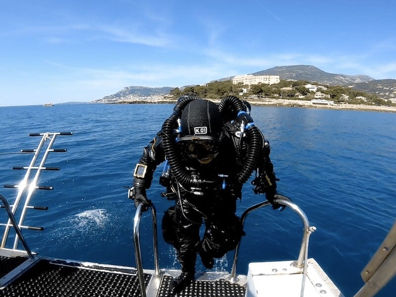 Plongeur en combinaison noire avec un équipement de respiration complexe remontant sur un bateau, avec la mer et une côte montagneuse en arrière-plan