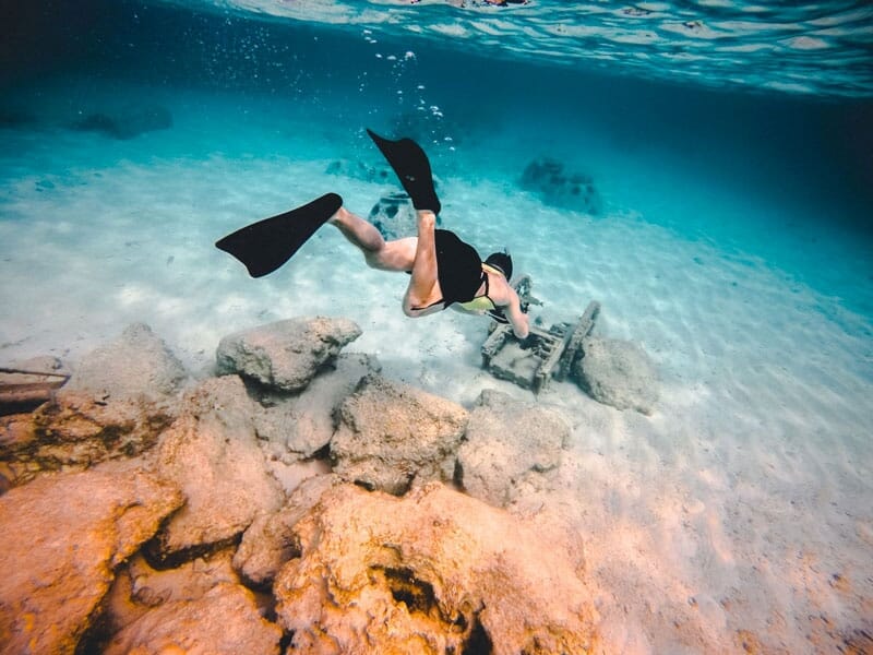 Plongeur en apnée avec palmes noires explorant un fond marin de sable blanc parsemé de rochers, dans une eau cristalline