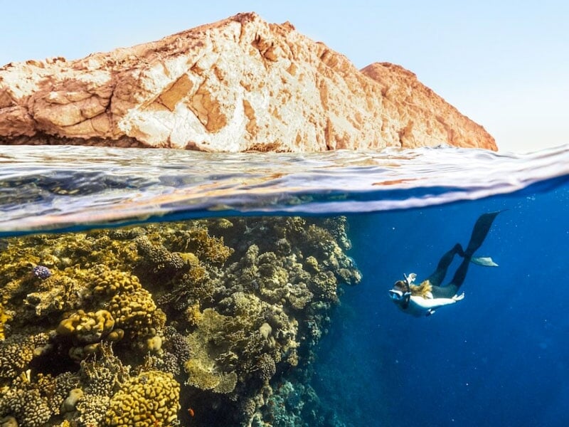 Vue mi-air mi-eau d'un plongeur en apnée nageant le long d'un récif corallien coloré, avec une falaise rocheuse en arrière-plan sous un ciel dégagé