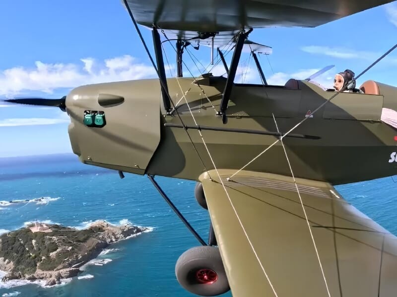 Avion biplan militaire vert olive en vol au-dessus de l'océan, avec un pilote portant des lunettes d'aviateur, ciel bleu parsemé de nuages blancs.