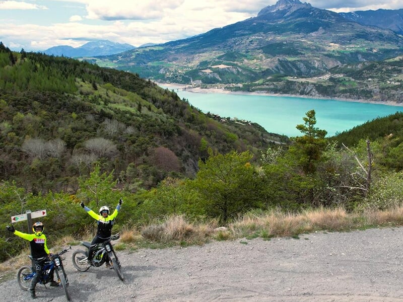 Des motards lèvent les bras de joie sur un chemin de terre avec le lac de Serre-Ponçon en toile de fond.