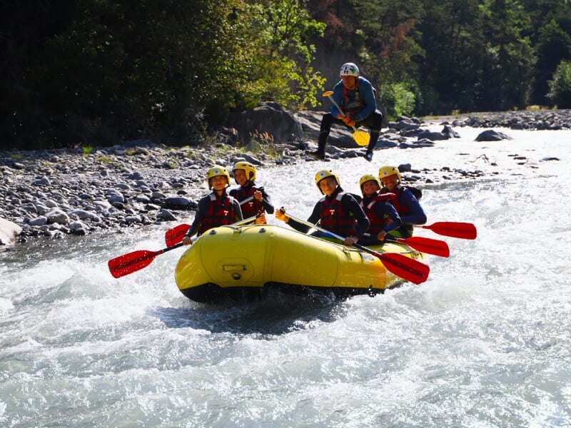 Groupe de six personnes en rafting sur une rivière rapide, pagayant dans un radeau jaune, portant des casques et des gilets de sauvetage, avec un guide debout à l'arrière du radeau. L'action se déroul