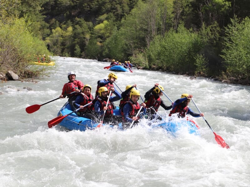 Équipe de huit personnes en rafting, pagayant dans un radeau bleu sur une rivière tumultueuse. Tous portent des casques jaunes et des gilets de sauvetage rouges. Des arbres verdoyants bordent la riviè