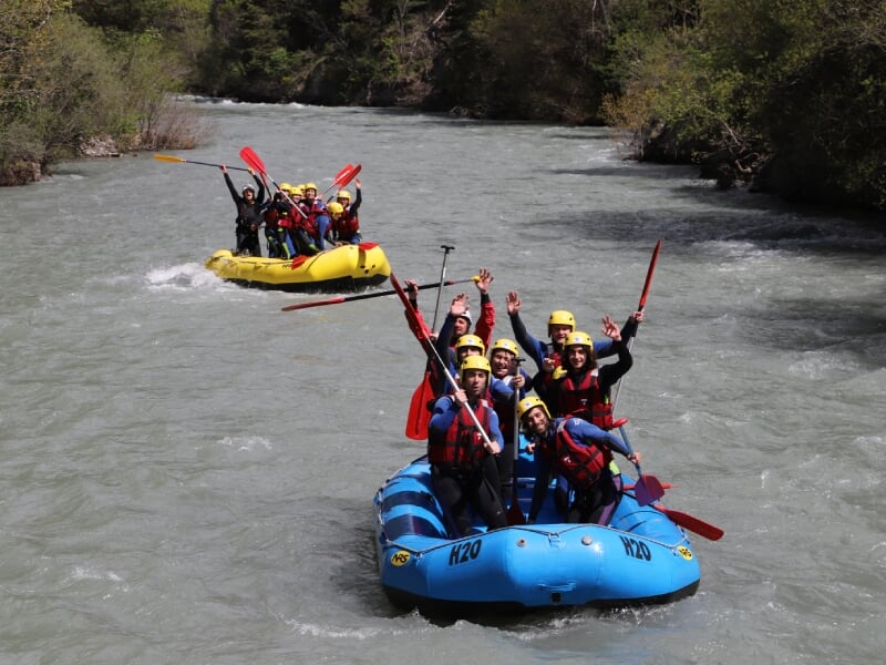 Deux groupes de personnes en rafting, un dans un radeau bleu au premier plan, l'autre dans un radeau jaune à l'arrière-plan, pagayant sur une rivière calme. Les participants lèvent les bras en signe d