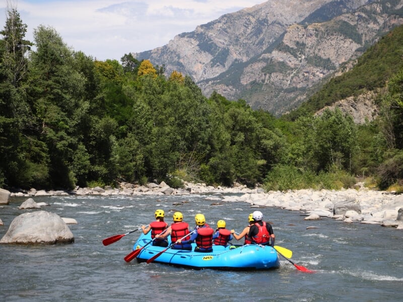 Groupe de six personnes pagayant dans un radeau bleu sur une rivière calme, entourée de montagnes et de végétation dense. Tous les membres du groupe portent des casques jaunes et des gilets de sauveta