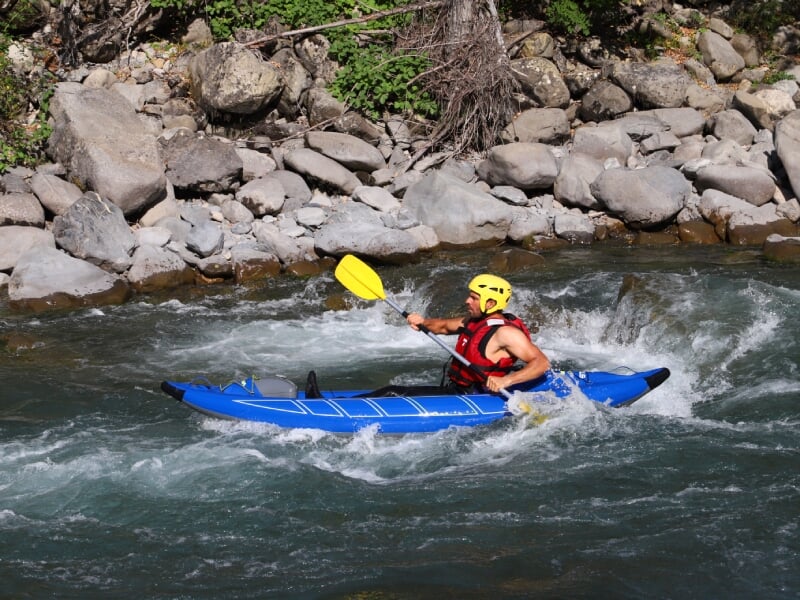 Homme pagayant en kayak bleu sur une rivière rapide, concentré, portant un casque jaune et un gilet de sauvetage rouge, avec des rochers et des arbres en arrière-plan.