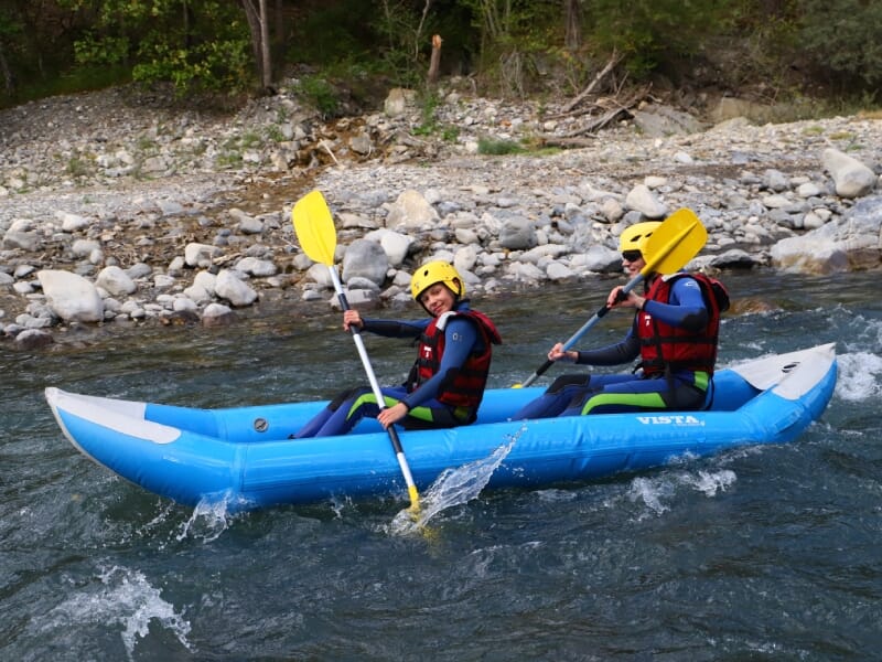 Deux personnes en kayak gonflable bleu sur une rivière calme, pagayant ensemble, portant des casques jaunes et des gilets de sauvetage rouges, avec une rive rocheuse en arrière-plan.