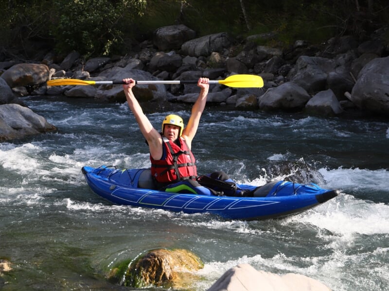 Homme en kayak bleu sur une rivière tumultueuse, pagayant avec enthousiasme, portant un casque jaune et un gilet de sauvetage rouge, entouré de rochers et de végétation.