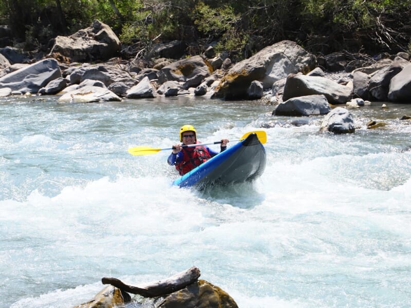 Homme en kayak bleu sur une rivière tumultueuse, pagayant avec force, portant un casque jaune et un gilet de sauvetage rouge, avec des rochers et une souche d'arbre visible.