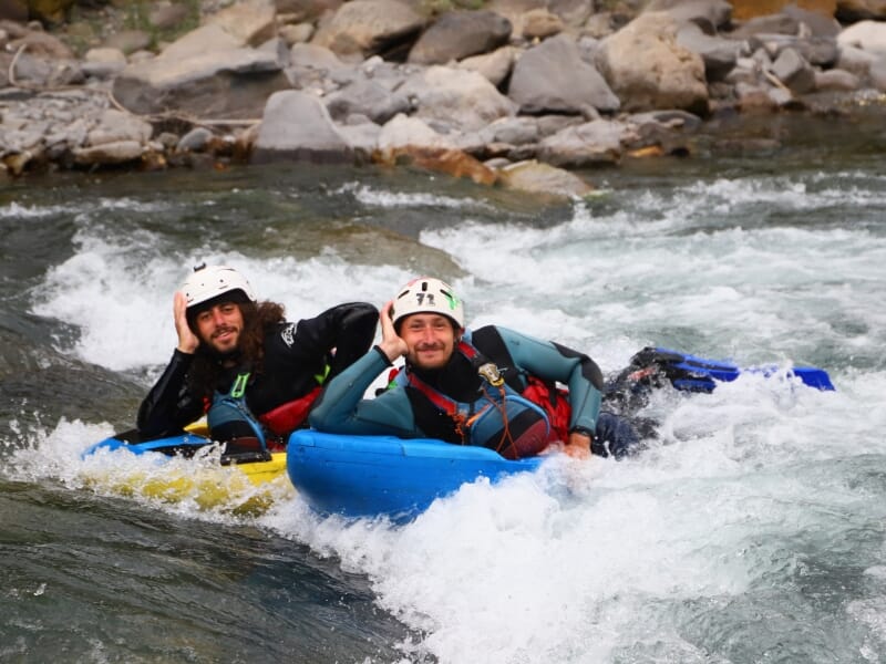 Deux hommes en hydrospeed, l'un sur une planche jaune et l'autre sur une bleue, souriant et saluant tout en descendant une rivière tumultueuse.