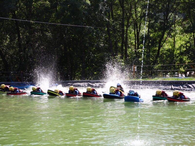 Groupe de personnes en hydrospeed alignées dans une piscine, éclaboussant l'eau en avançant, entourées d'arbres et de verdure.