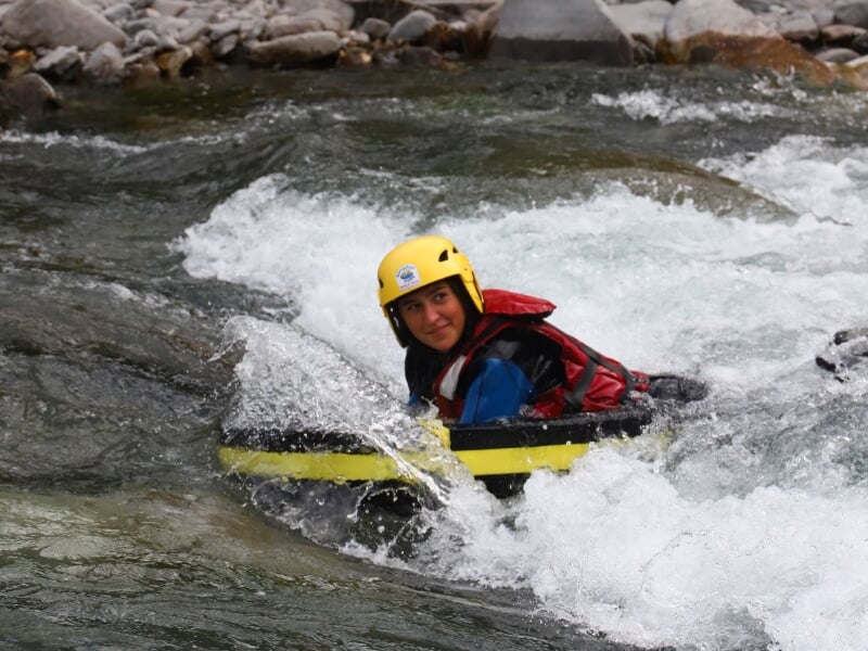 Jeune femme en hydrospeed avec un casque jaune et une planche noire et jaune, naviguant dans une rivière avec de petites vagues et des rochers.