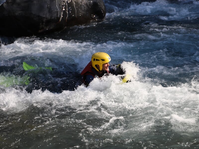 Pratiquant de hydrospeed portant un casque jaune et un gilet de sauvetage rouge, traversant une rapide avec de l'eau éclaboussante autour.