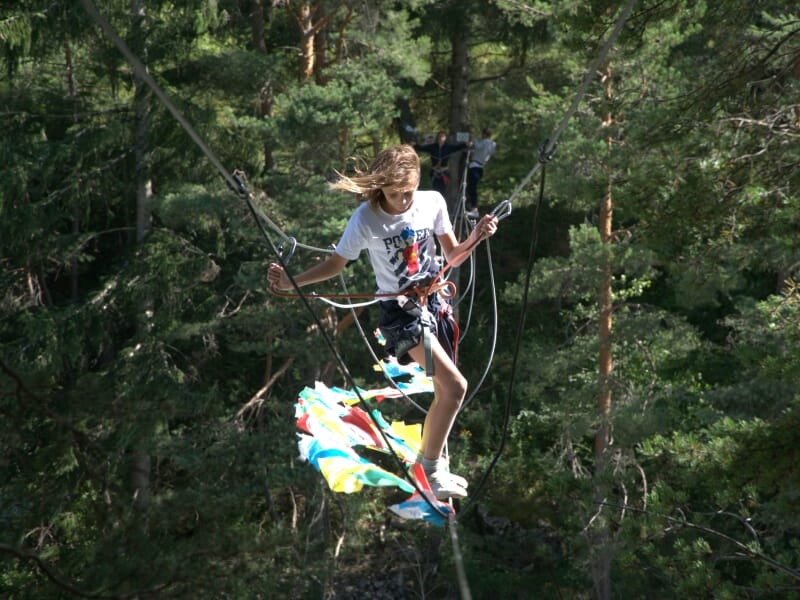 Adolescente sur une passerelle suspendue colorée, équipée d'un harnais de sécurité, cheveux au vent, forêt dense en arrière-plan.