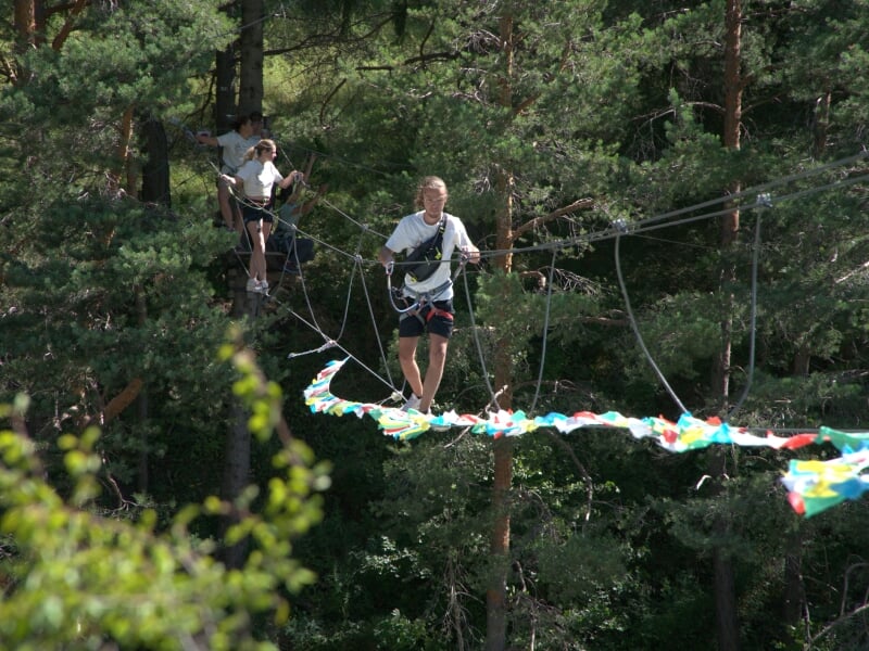 Homme marchant sur une passerelle suspendue dans les arbres, portant un harnais de sécurité et des vêtements de sport, forêt dense en arrière-plan.