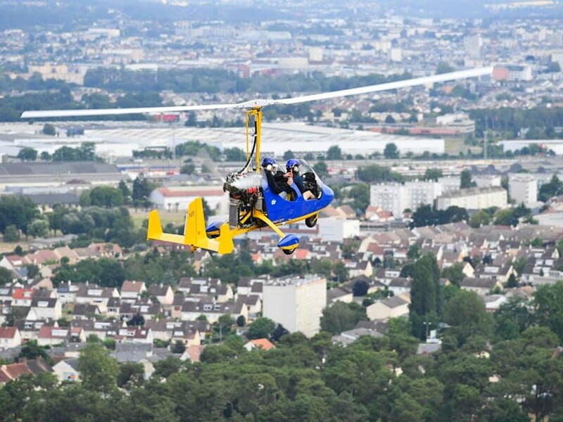 ULM bleu en vol avec deux passagers portant casque, survolant une ville et des collines.