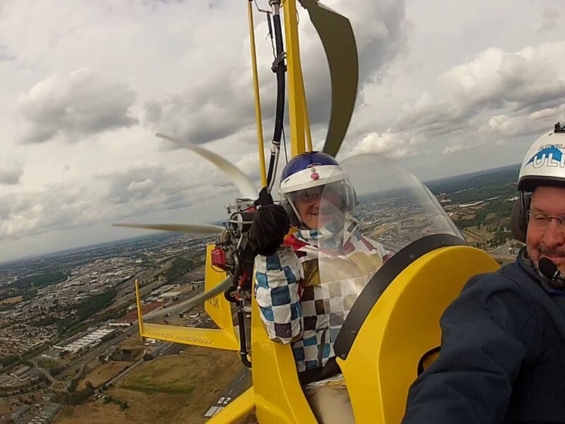 Deux personnes dans un ULM jaune en vol, l'une faisant un signe de la main, ciel nuageux.