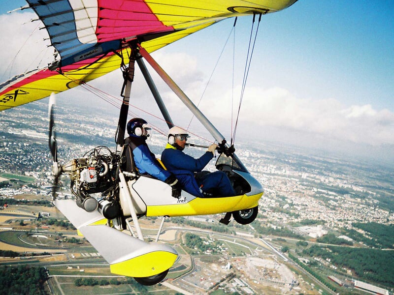 ULM jaune en vol avec deux passagers portant casque, survolant une ville et ses environs.