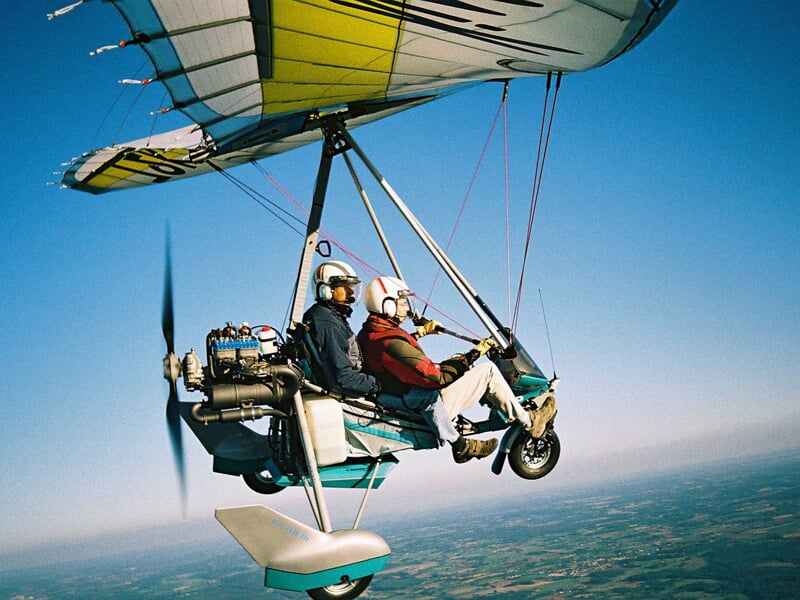 ULM en vol avec deux personnes équipées de casques, survolant un paysage rural sous un ciel bleu.